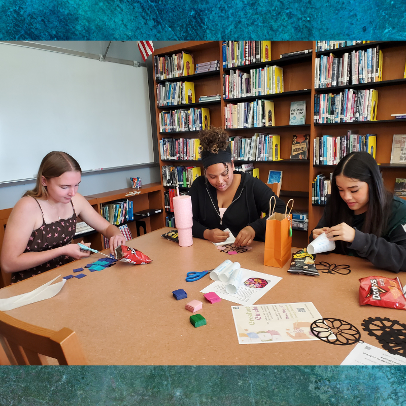 Three young people are engaged in a craft activity at a table in a library, with bookshelves in the background.