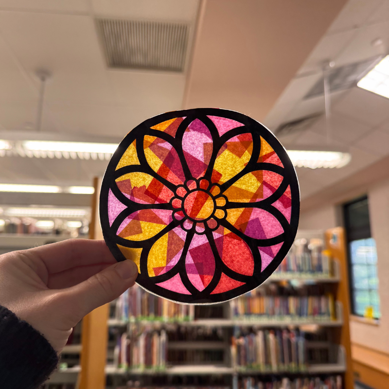 A hand holds a colorful, circular stained-glass-like craft in front of library bookshelves.
