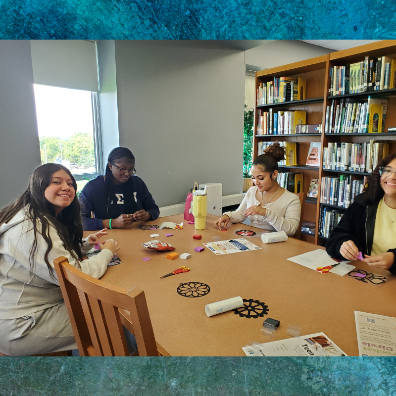Four young people are engaged in a craft activity at a table in a library, with bookshelves in the background.