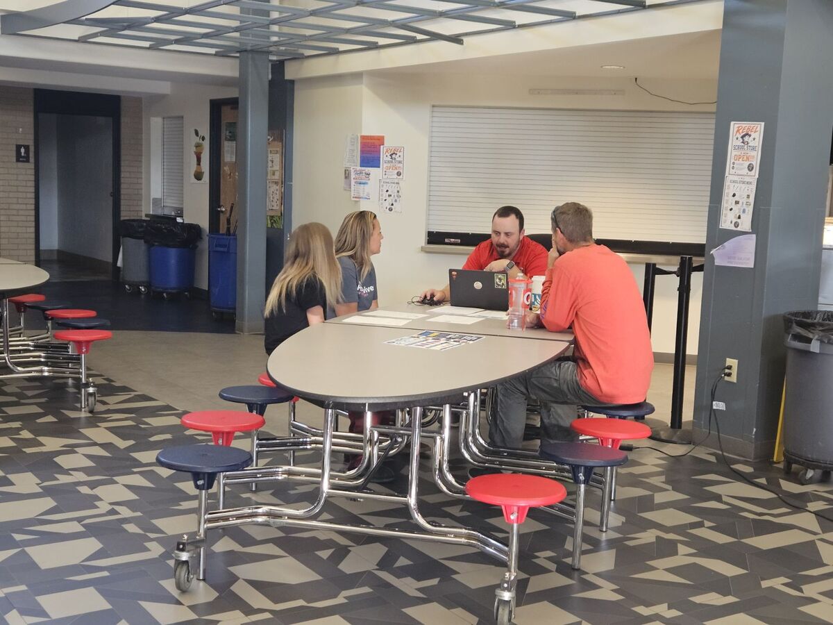Four people sit around a cafeteria table, collaborating with a laptop in a school hallway.