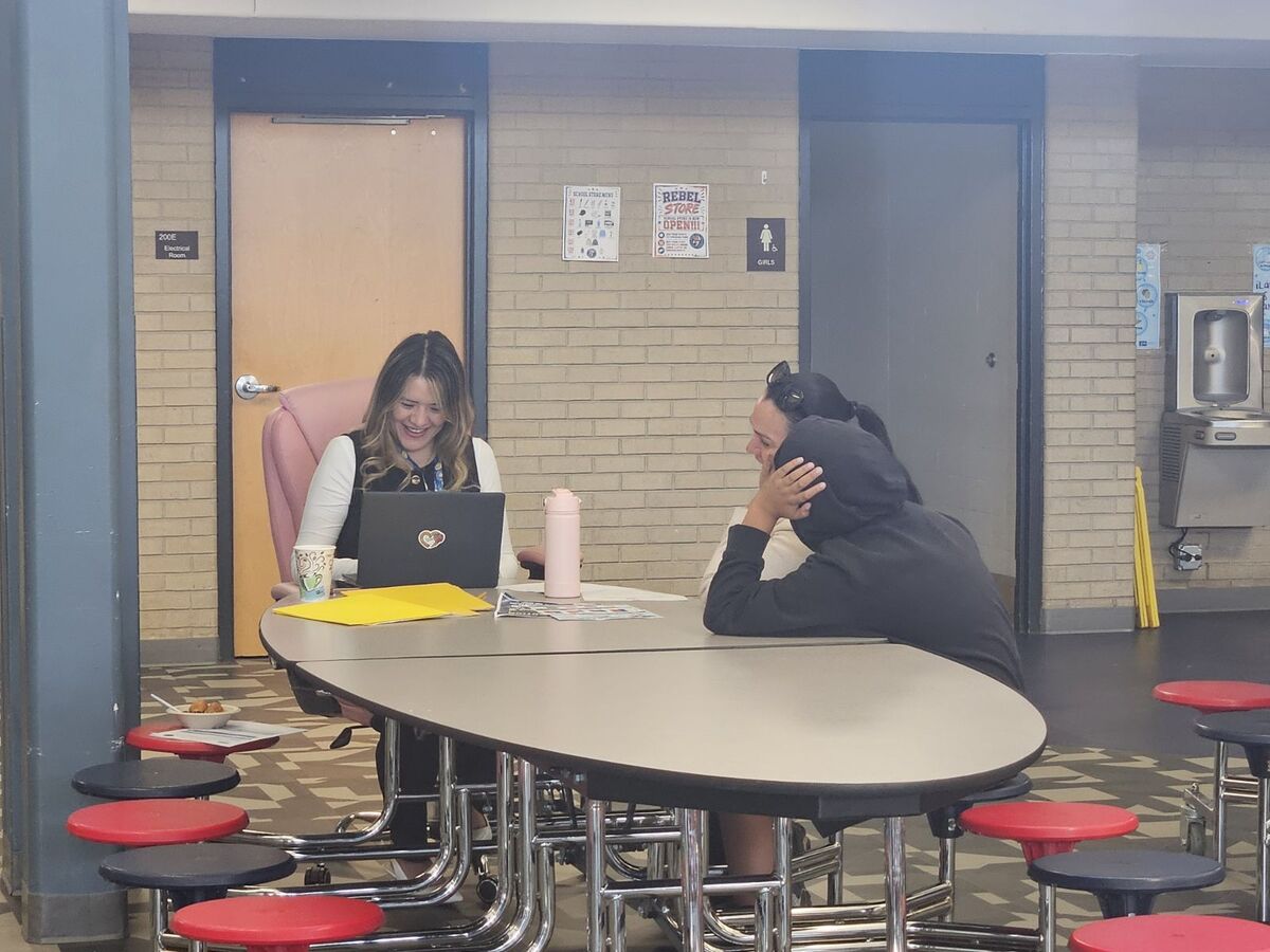 A woman smiles while using a laptop at a table in a school hallway with a student.