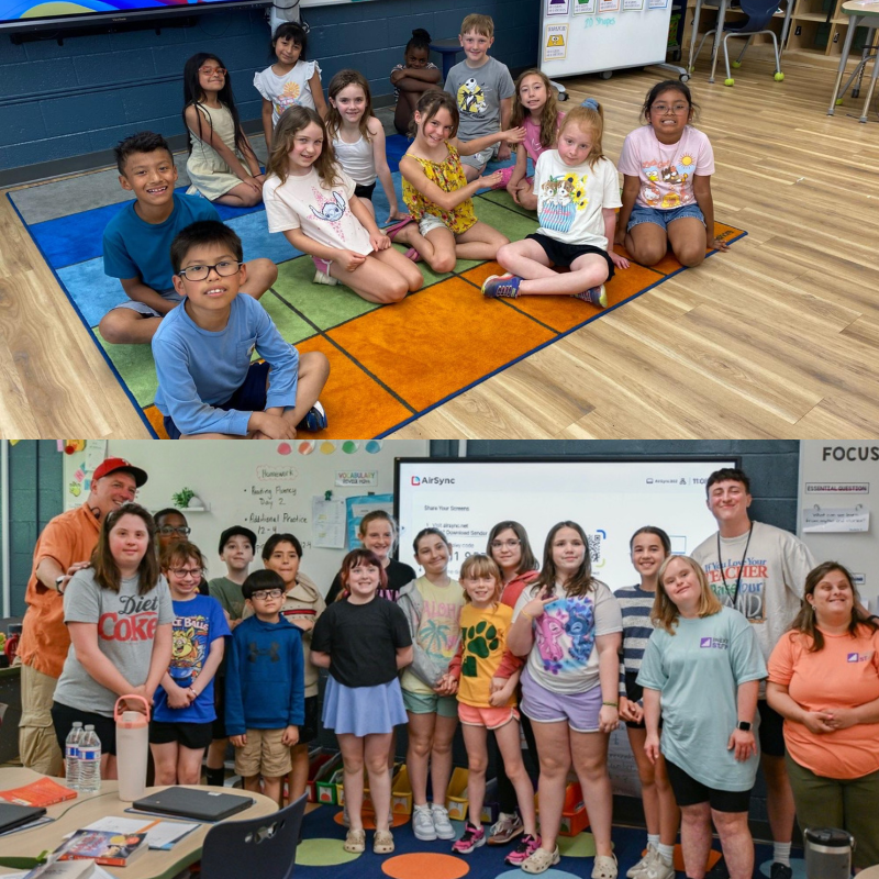 A group of children and two adults pose for a photo in a classroom, with a whiteboard and projector screen behind them.