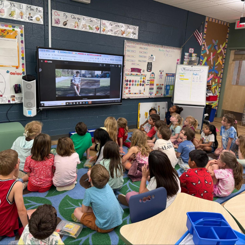 A diverse group of young students sit on a colorful rug, attentively watching a video on a large screen in a classroom.
