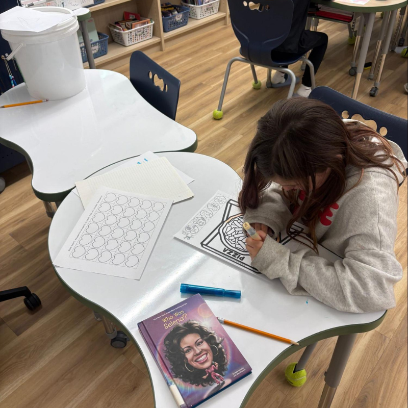 A young girl colors a "Pizza" drawing at a white table in a classroom.