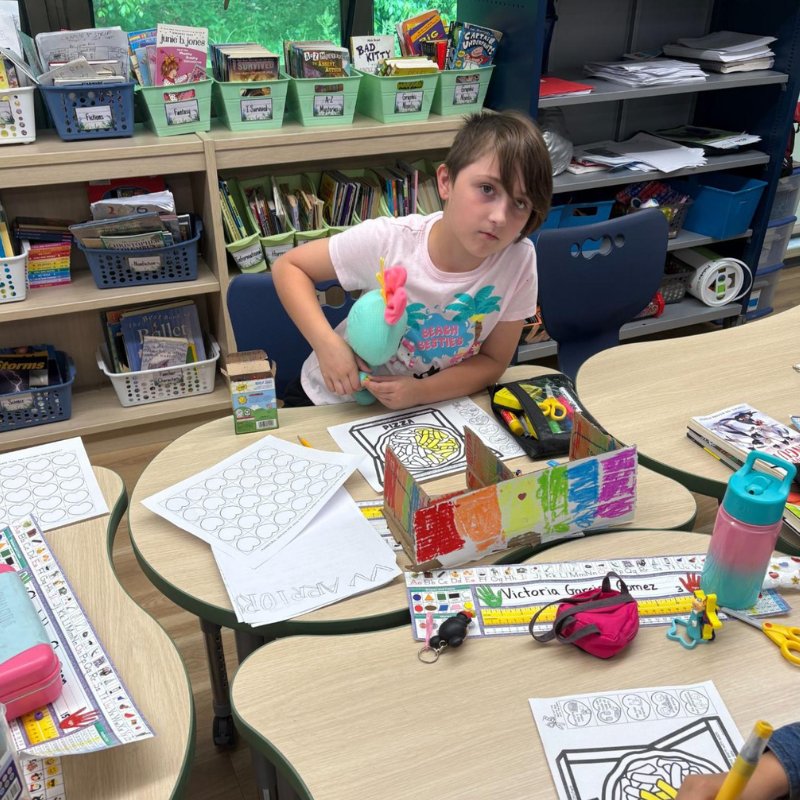 A young student sits at a classroom table, holding a stuffed toy, with art supplies and coloring pages spread out.
