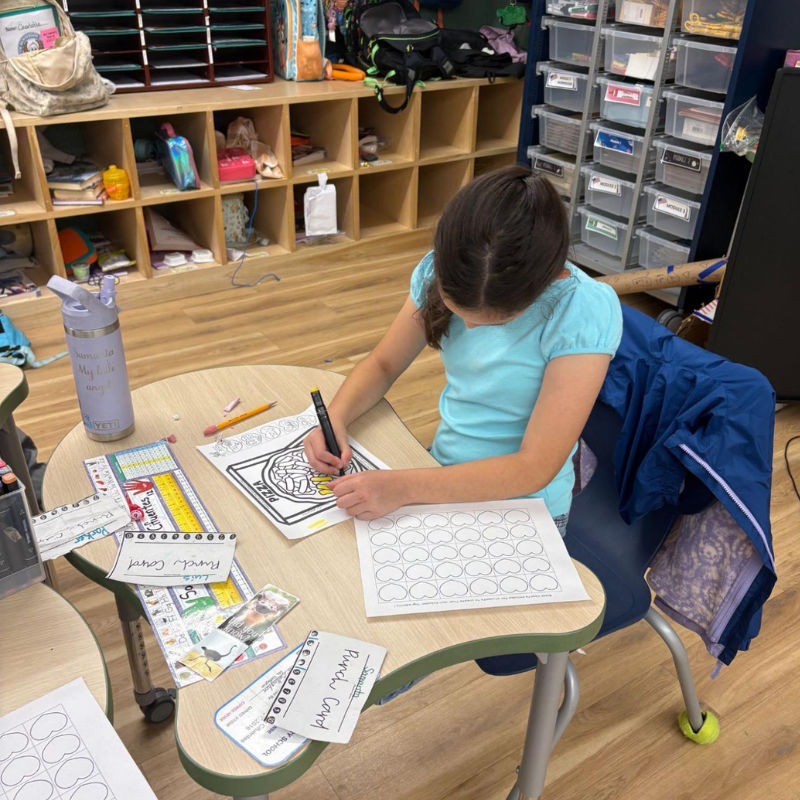 A young girl in a blue shirt colors a pizza drawing at a classroom desk.