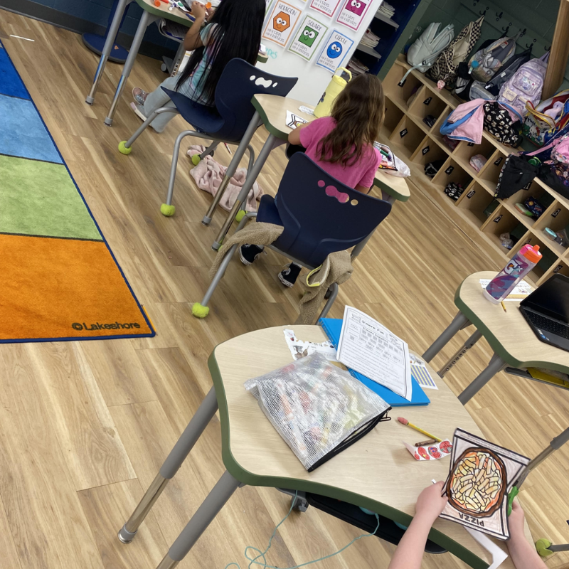 Children work at desks in a classroom, with colorful rugs and cubbies visible.