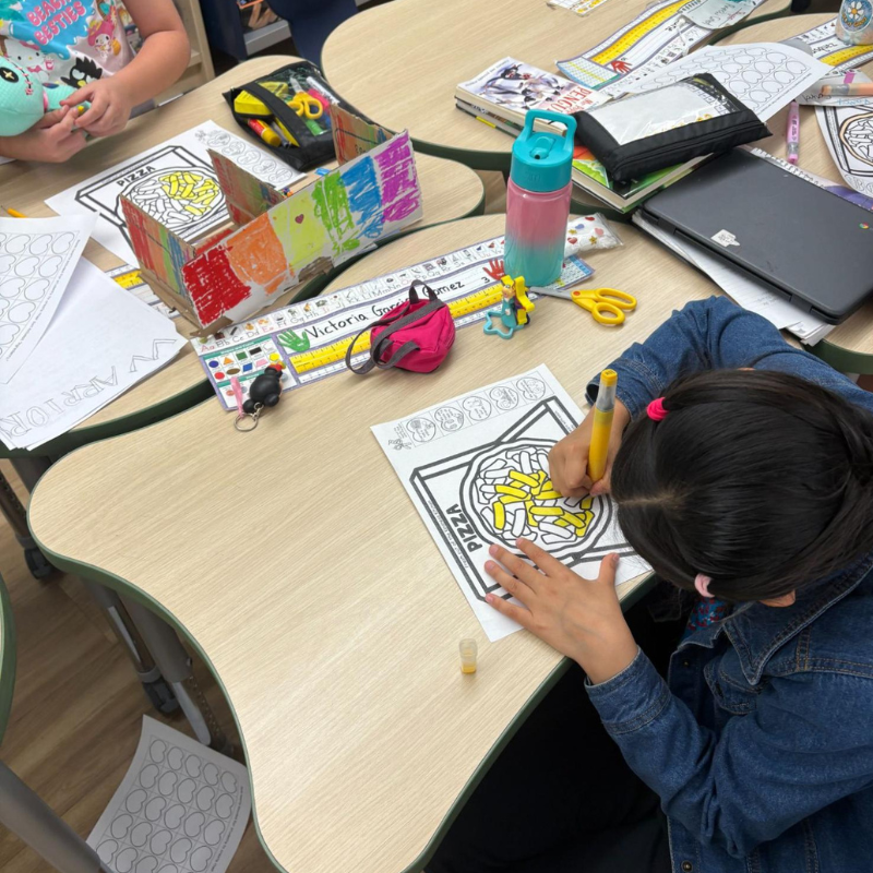 A young student colors a pizza-themed worksheet at a classroom desk.