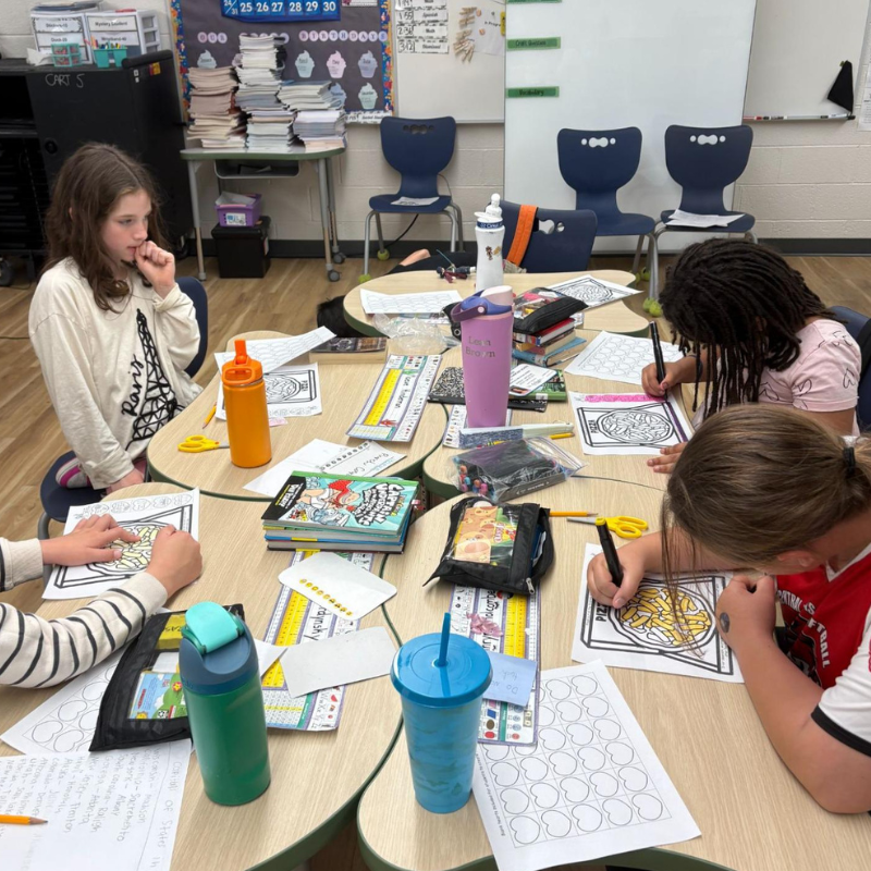 Students are coloring at round tables in a classroom, with art supplies and books spread out.