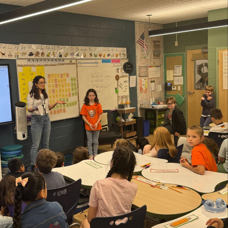 A teacher and a student stand near a whiteboard in a classroom, addressing a group of seated students.