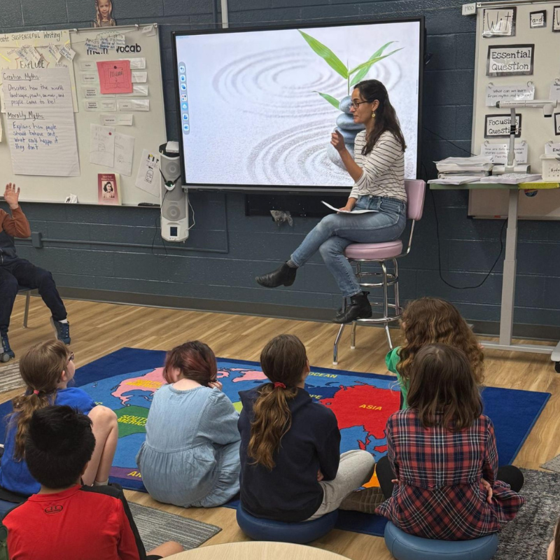 A teacher sits on a stool in front of a large screen displaying a zen garden image, engaging with students seated on a world map rug.