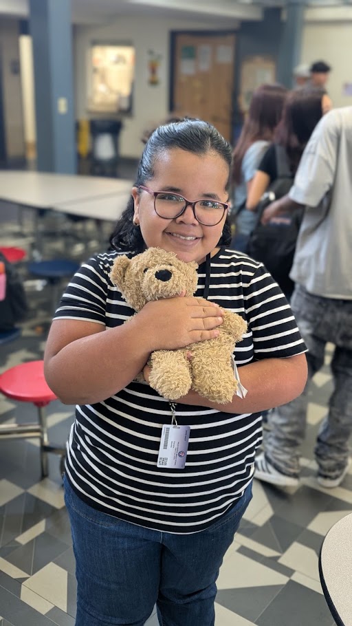 A smiling person wearing glasses and a striped shirt holds a small stuffed bear in a brightly lit room.