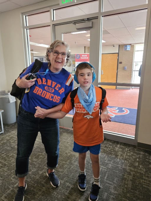 A smiling woman and a young boy stand together inside a building, both wearing Denver Broncos gear.