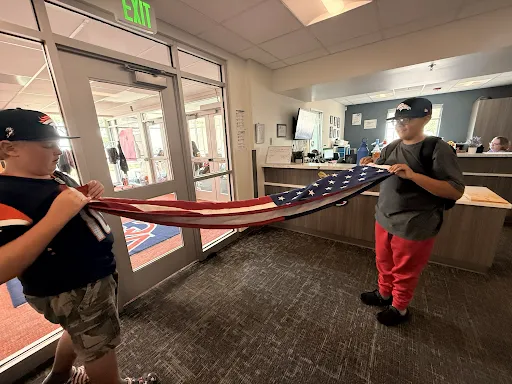 Two kids hold a folded American flag together inside a brightly lit building foyer.