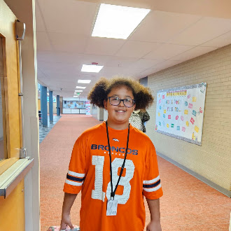 A smiling child wearing an orange Broncos jersey stands in a school hallway.