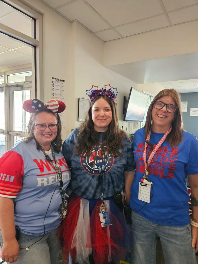 Three smiling adults stand indoors, dressed in festive attire with patriotic headbands and casual shirts.