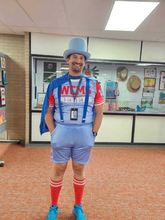 A smiling man in a blue top hat, matching cape, and jersey with the text "WMS REBELS" stands indoors.