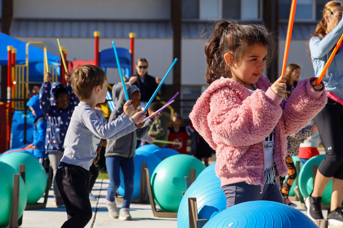 Children and adults participate in an outdoor music and fitness class with drumsticks and exercise balls.