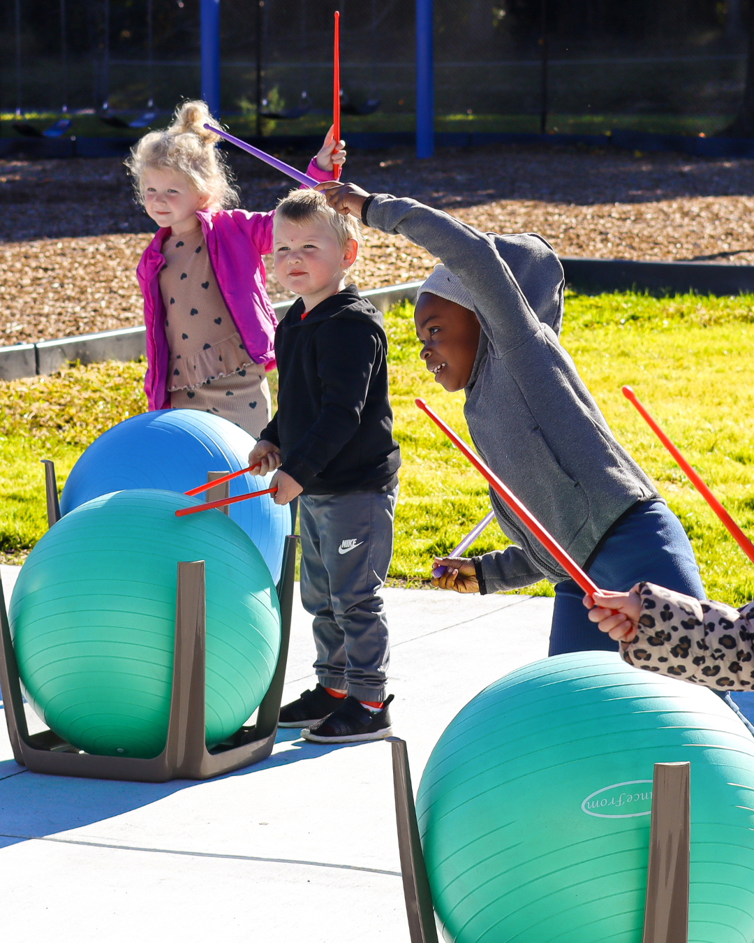 Three children joyfully play music outdoors, using drumsticks on large exercise balls held in stands.