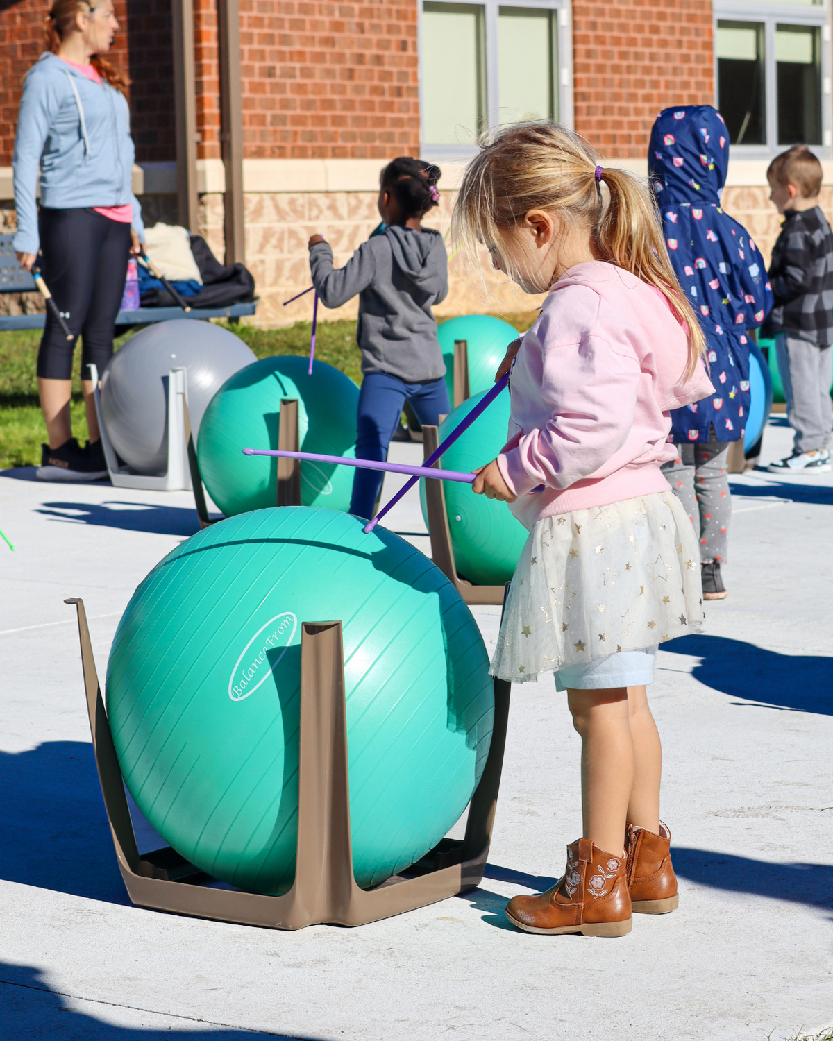A young child in a pink hoodie hits a teal exercise ball with drumsticks outdoors.