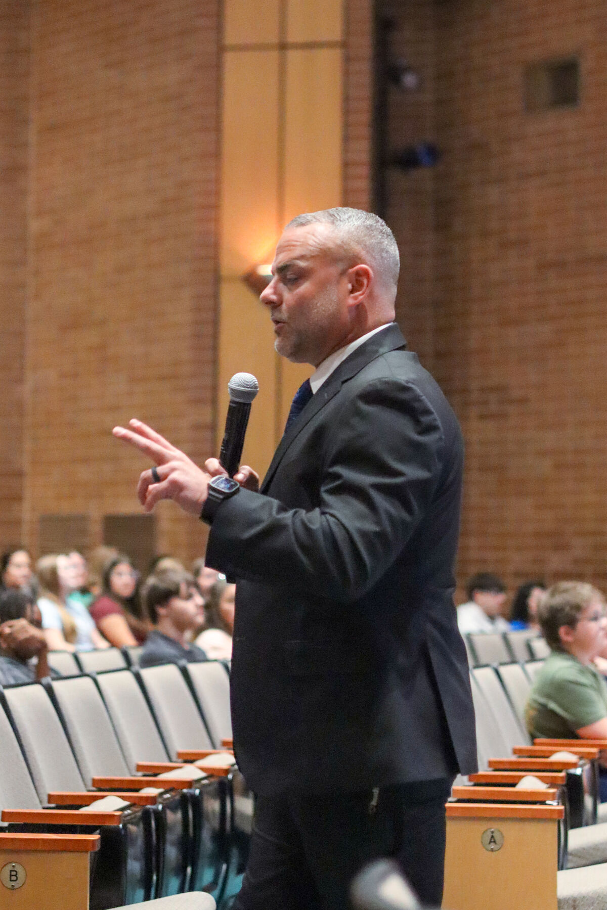 A man in a suit speaks into a microphone while gesturing with his hands to an audience seated in an auditorium.