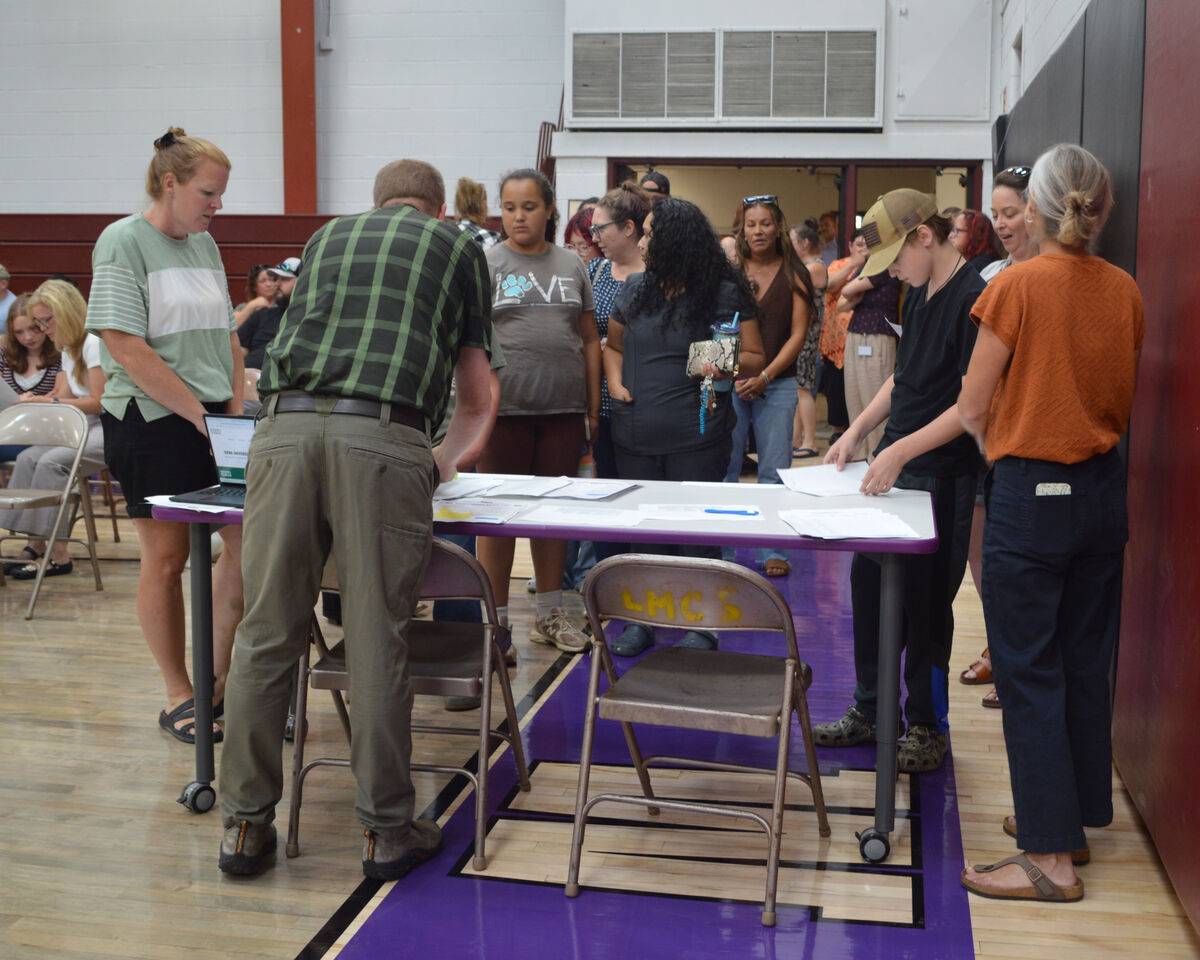 People gather around tables with papers and a laptop in a gymnasium.
