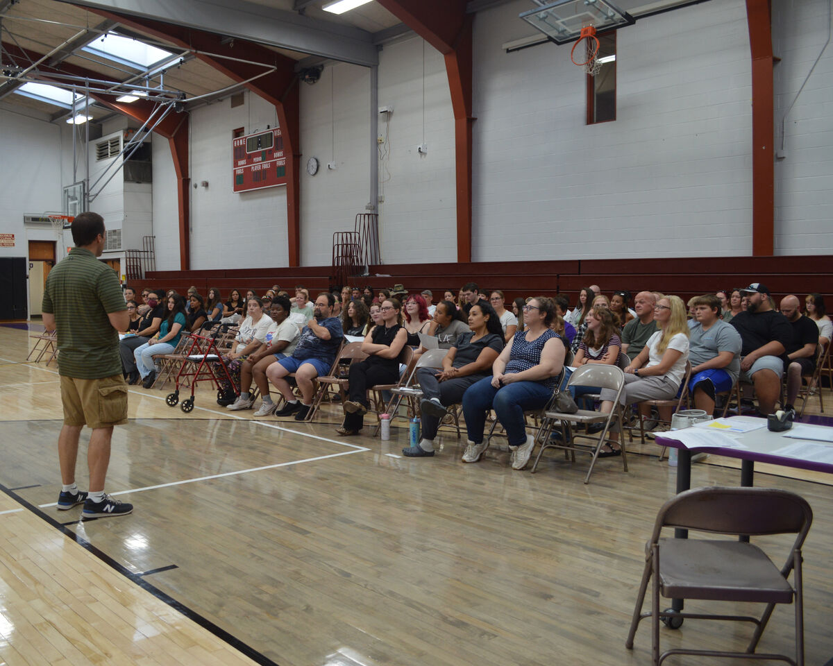 A man in a green striped shirt and khaki shorts stands facing an audience seated in a gymnasium.