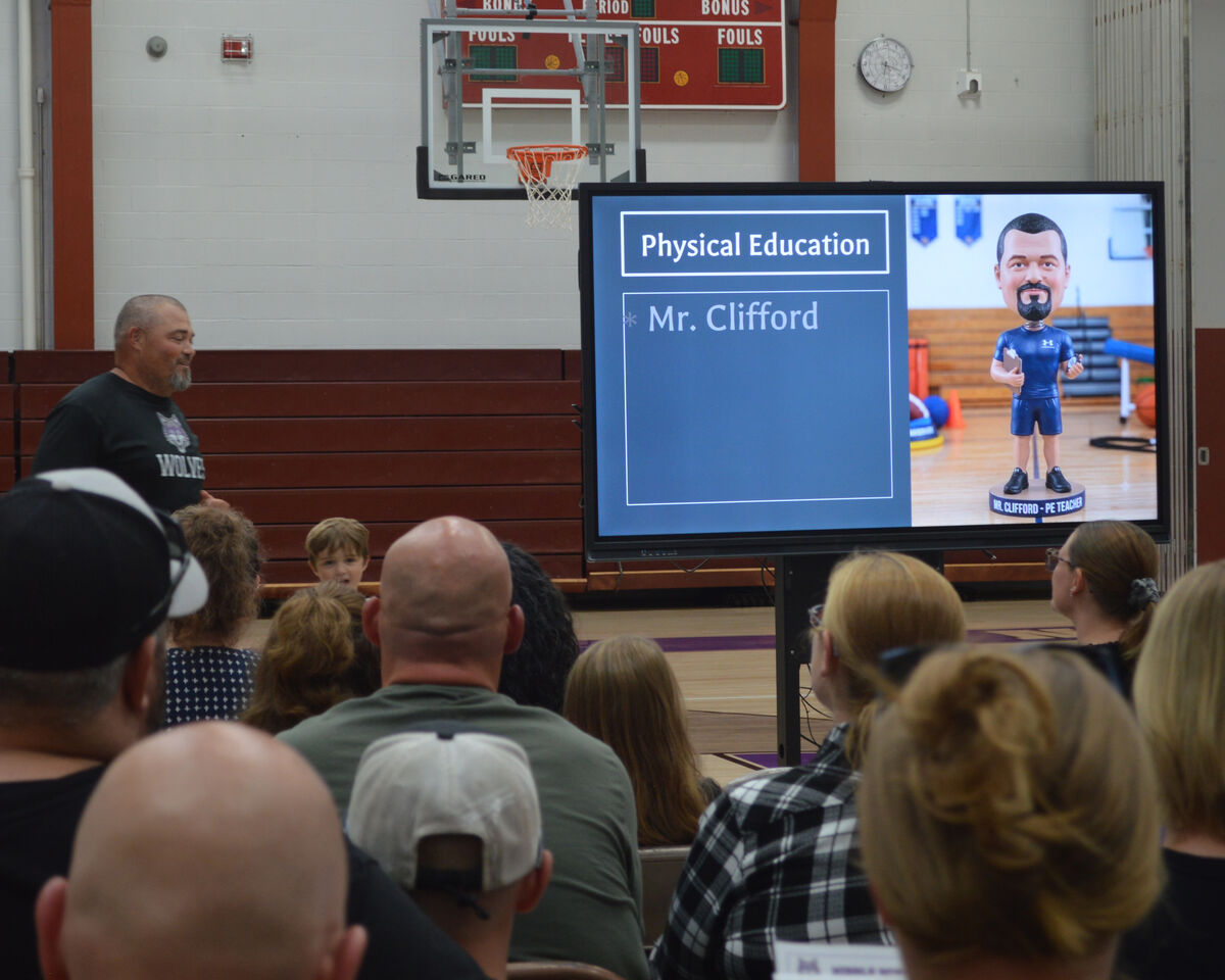 A man in a black t-shirt stands in front of a large screen displaying 'Physical Education' and 'Mr. Clifford'.
