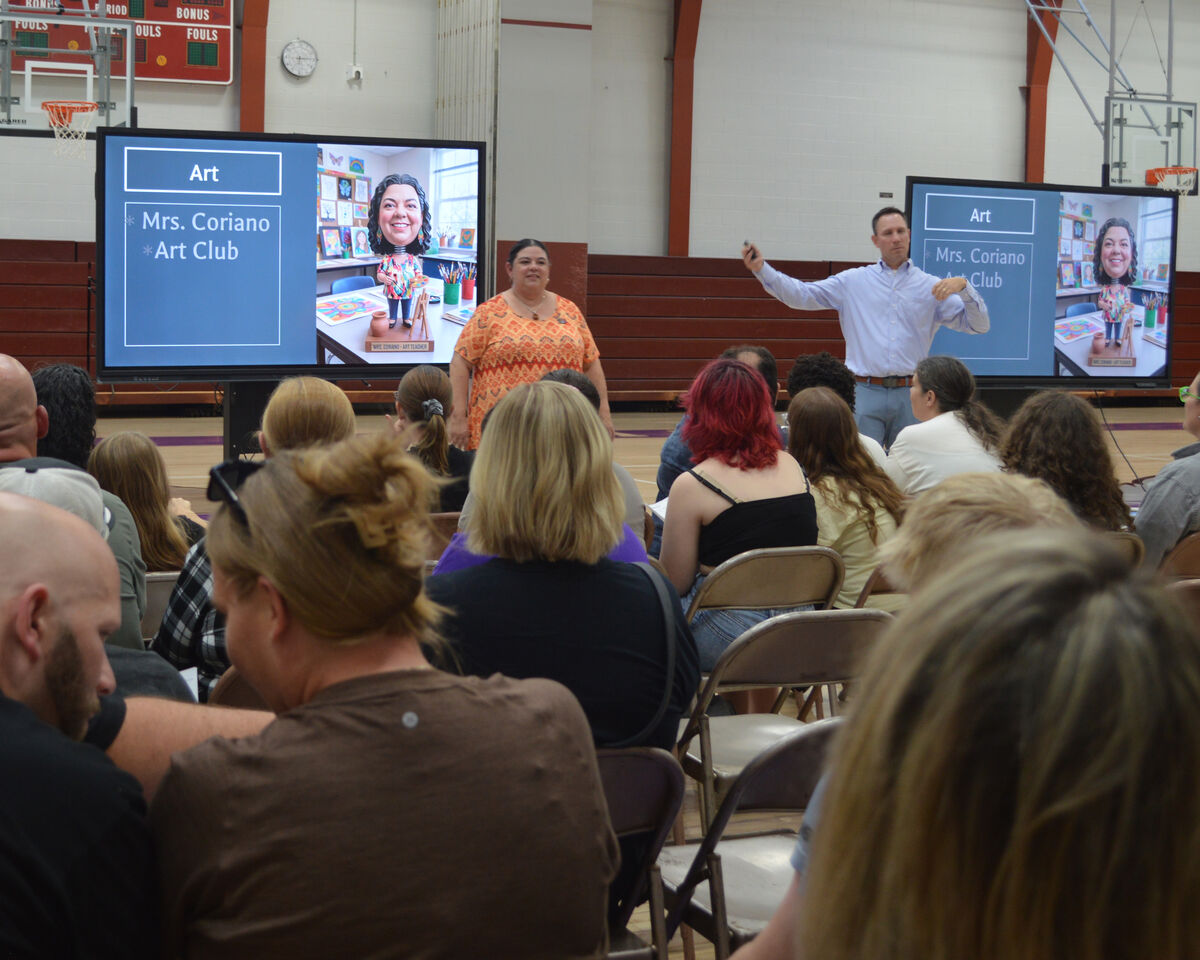 A man and a woman stand near large screens displaying 'Art' and 'Mrs. Coriano Art Club' in a gymnasium.
