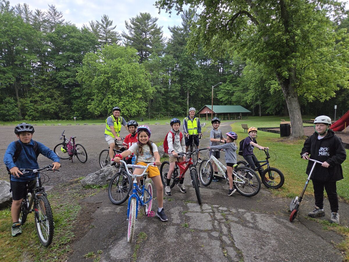 A group of children and adults in helmets stand with their bicycles in a park setting.