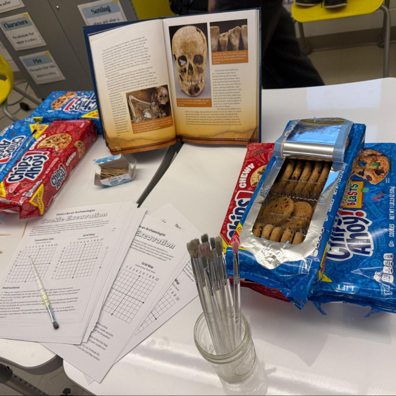An open book displays images of a skull and ancient artifacts, surrounded by cookie packages and excavation worksheets.
