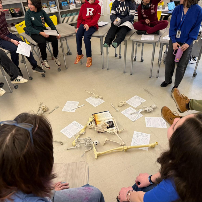 Students sit in a circle around a classroom floor with model skeletons and papers.
