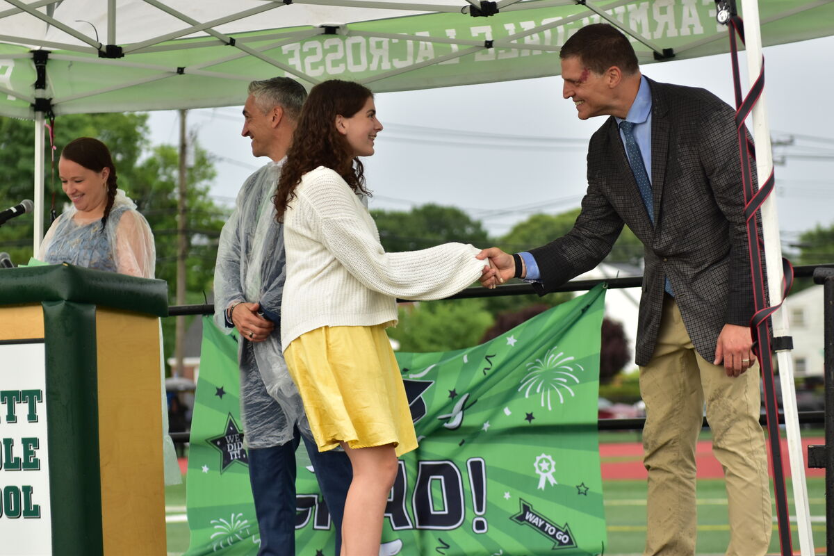 A young woman in a white sweater and yellow skirt shakes hands with a man in a suit on a stage.