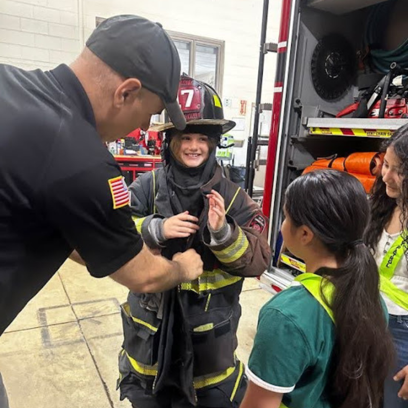 A firefighter helps a child try on a full firefighter's uniform and helmet.