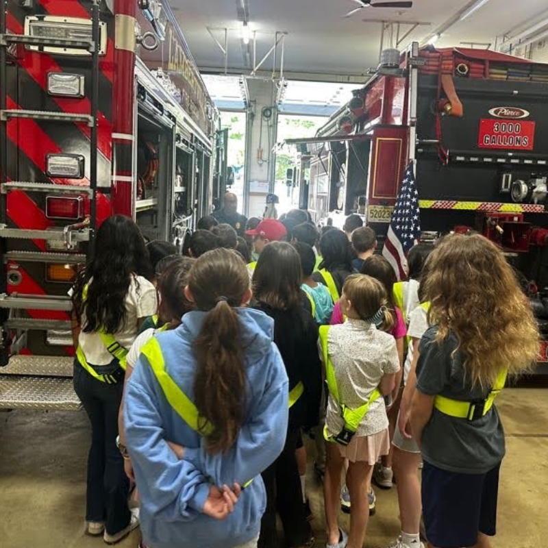 A group of children wearing safety vests stand in a fire station, looking at fire trucks.