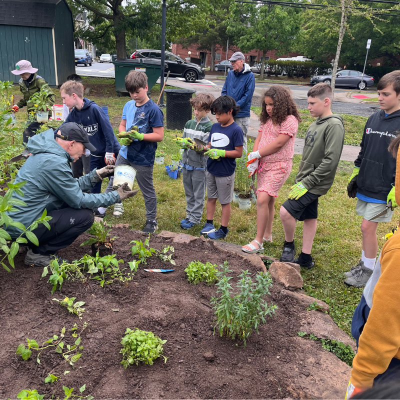 A group of children and adults are planting in a garden bed, with a man demonstrating how to use a bucket.