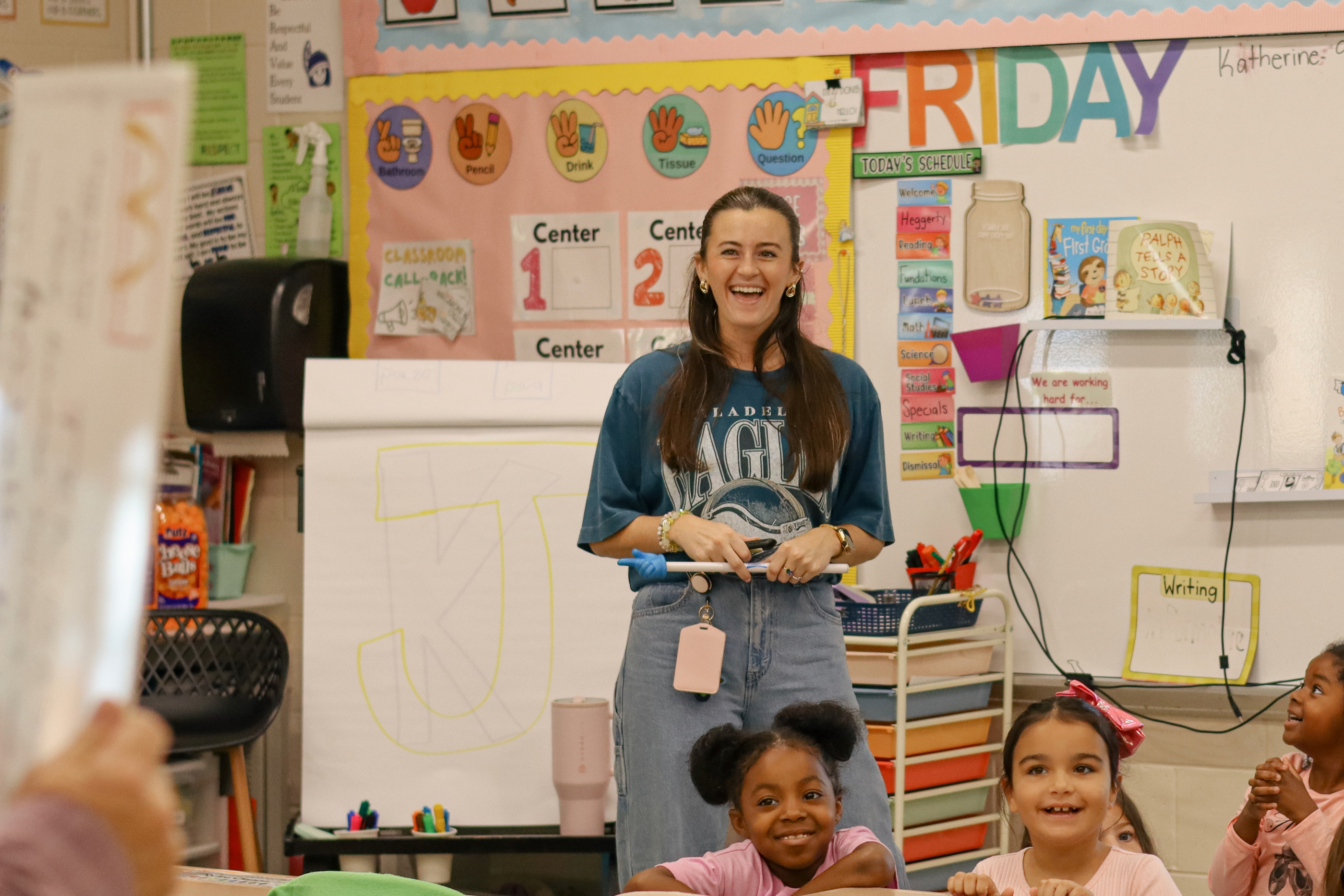 A teacher smiles brightly while standing in front of a colorful classroom with students.