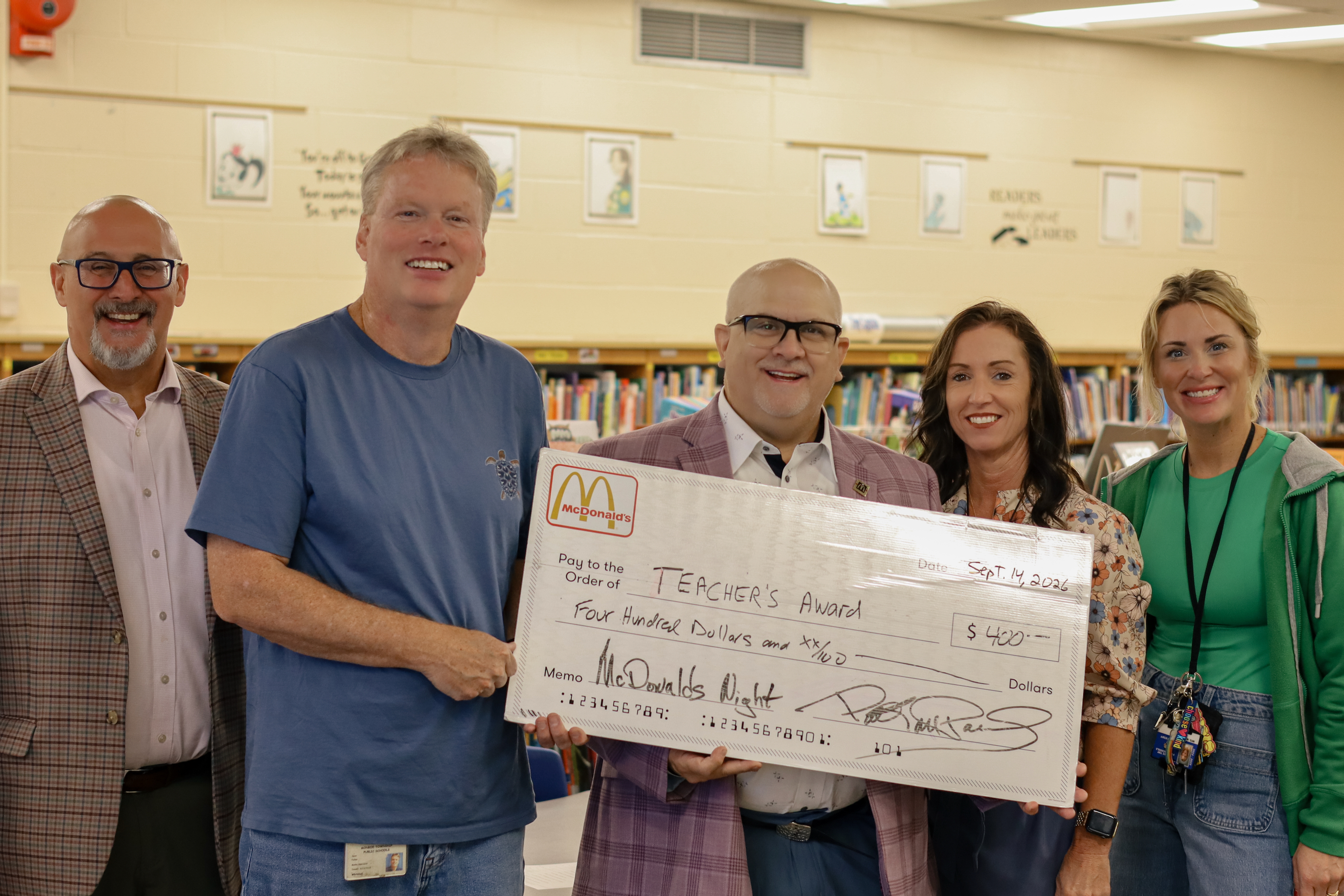 Five smiling adults proudly hold a large novelty check for a teacher's award inside a school library.
