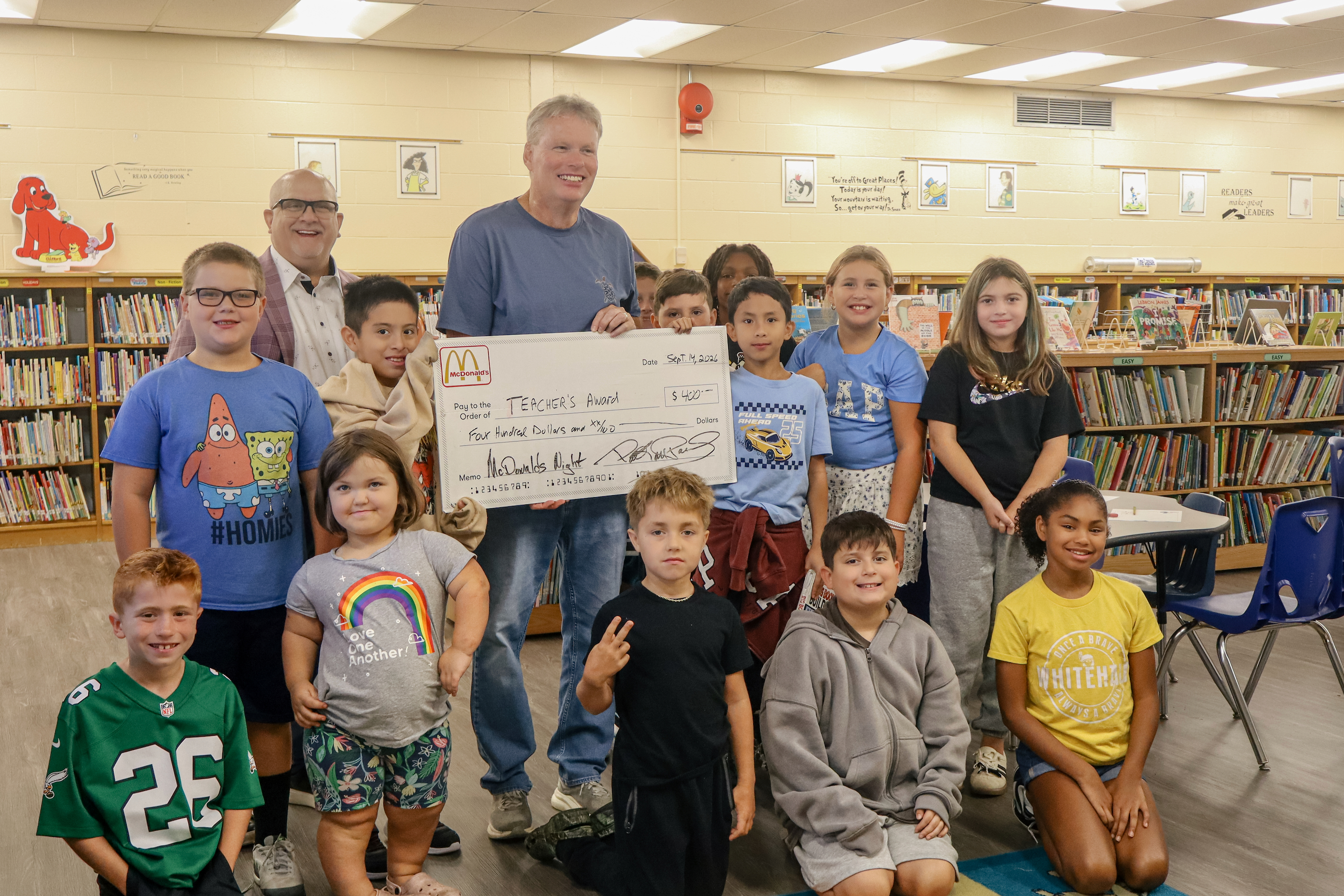 A smiling group of children and adults hold a large novelty check in a school library.