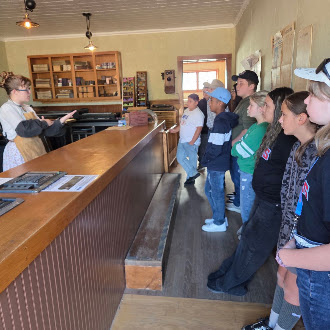 A guide in historical attire speaks to a group of visitors gathered inside a rustic shop with a wooden counter.