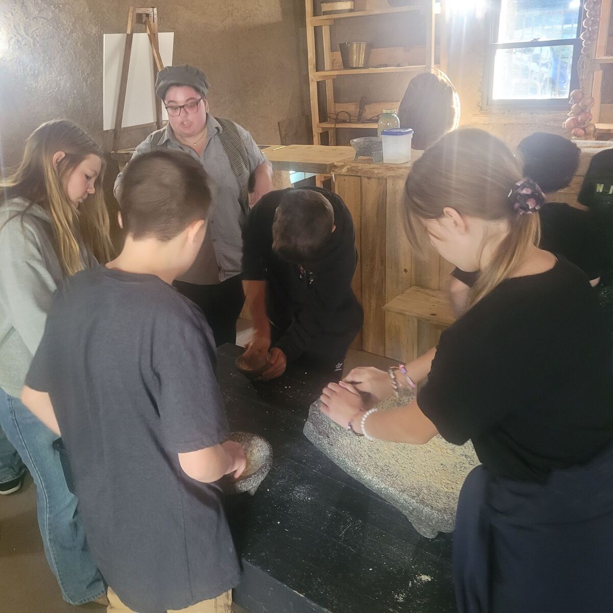 A group of students stands around a dark table, working with traditional stone grinding tools indoors.
