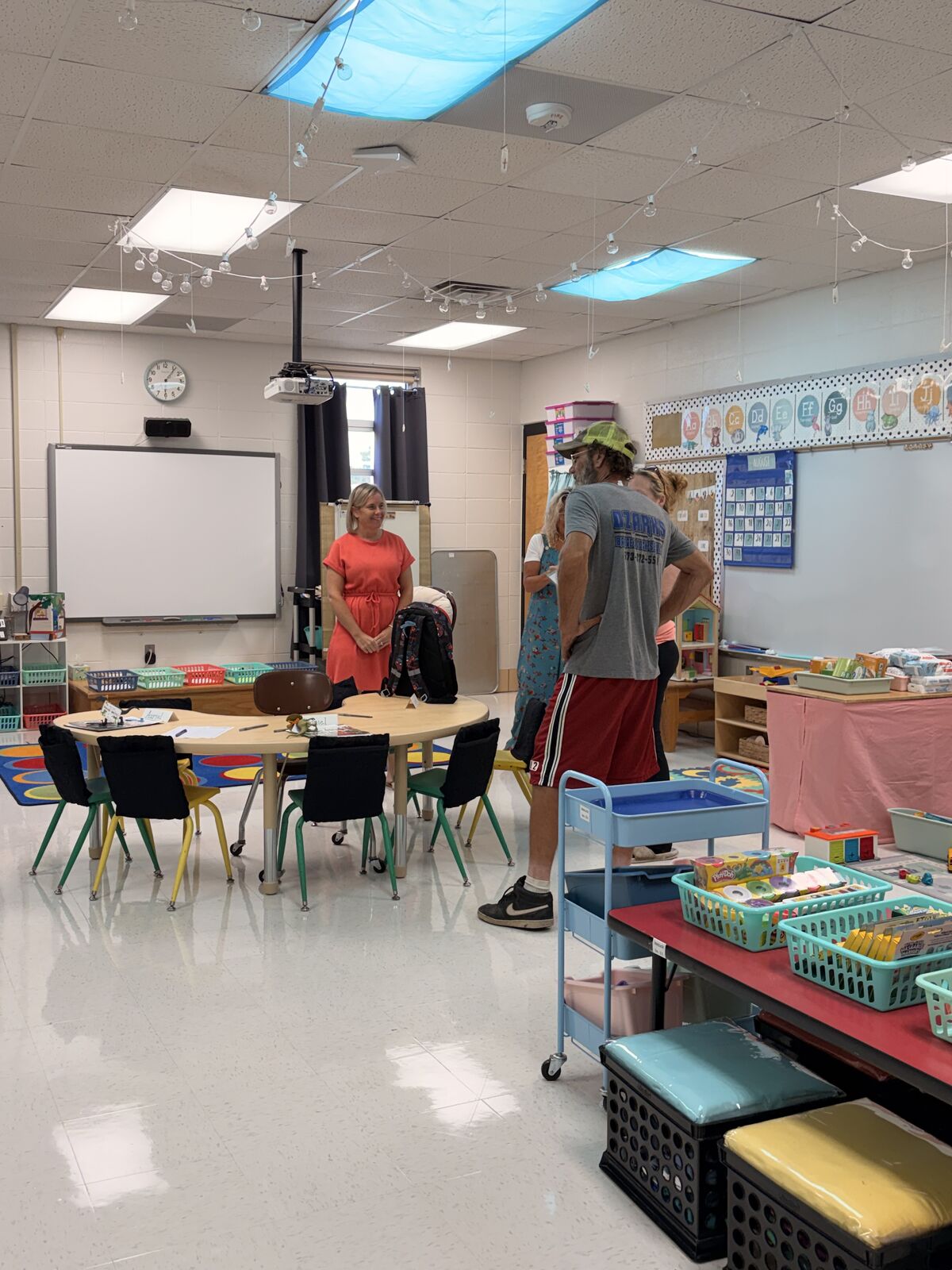 Adults in a classroom setting, with colorful baskets of supplies on shelves and a projector mounted on the ceiling.