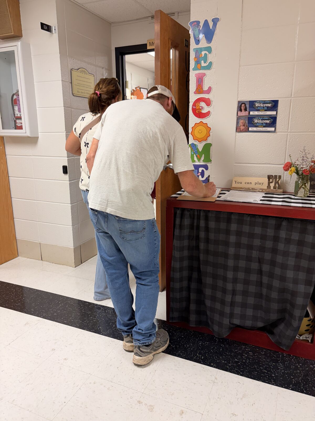 A man in a white t-shirt and jeans writes on a clipboard near a colorful "WELCOME" banner.