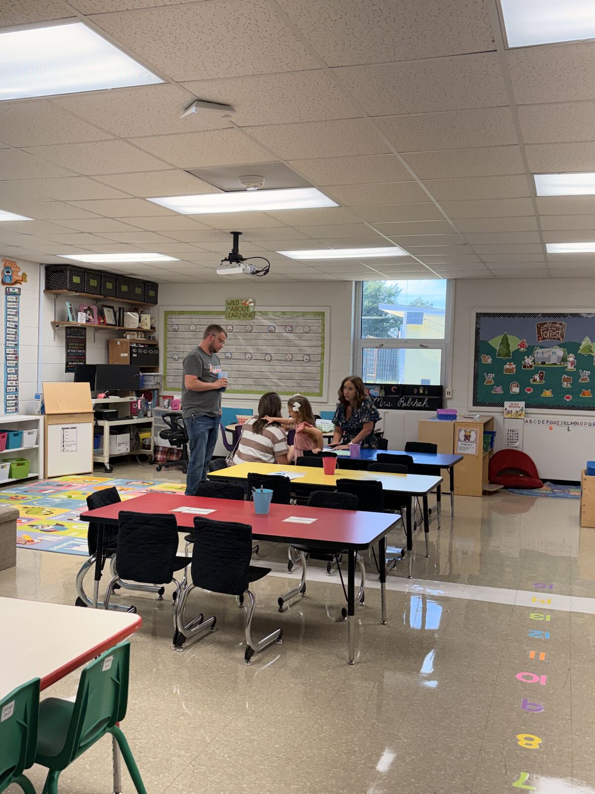 A teacher stands in front of a classroom of young children seated at colorful tables.