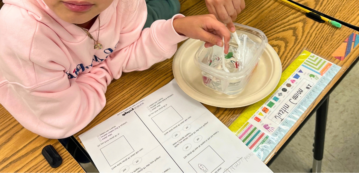 A student in a pink hoodie conducts a science experiment at a wooden desk.