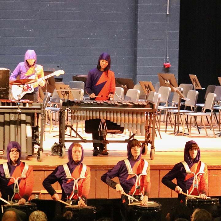A marching band performs on stage, with members playing drums, a guitar, and a xylophone.