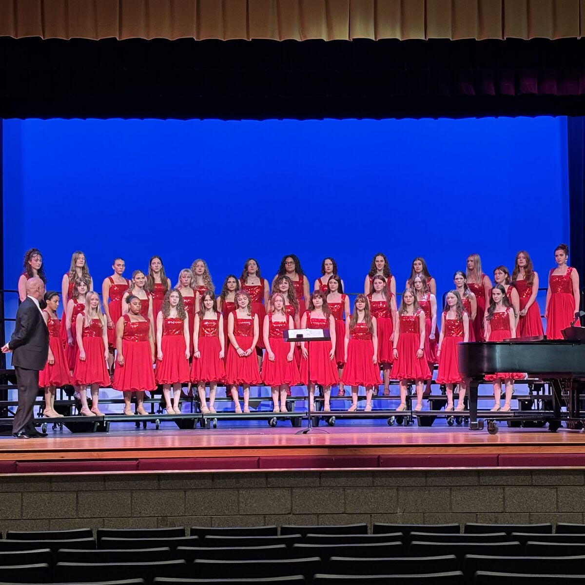 A choir of young women in red dresses stands on risers on a stage with a blue backdrop.