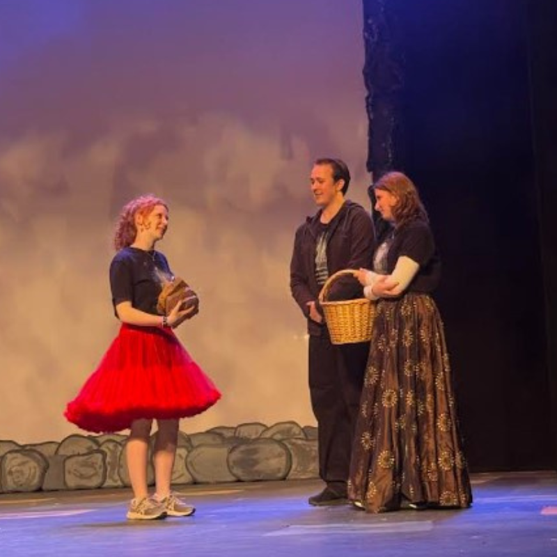 Three actors on a stage, one in a red skirt holding a basket, and two others holding a basket.