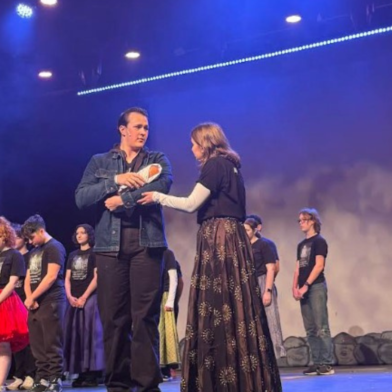 A young man in a denim jacket holds a prop baby while a young woman in a patterned skirt looks on during a stage performance.