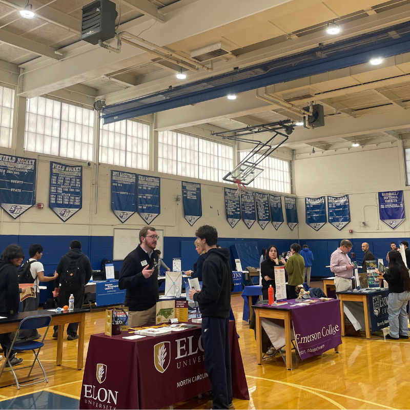 People interact at tables representing universities in a gymnasium with sports banners on the wall.